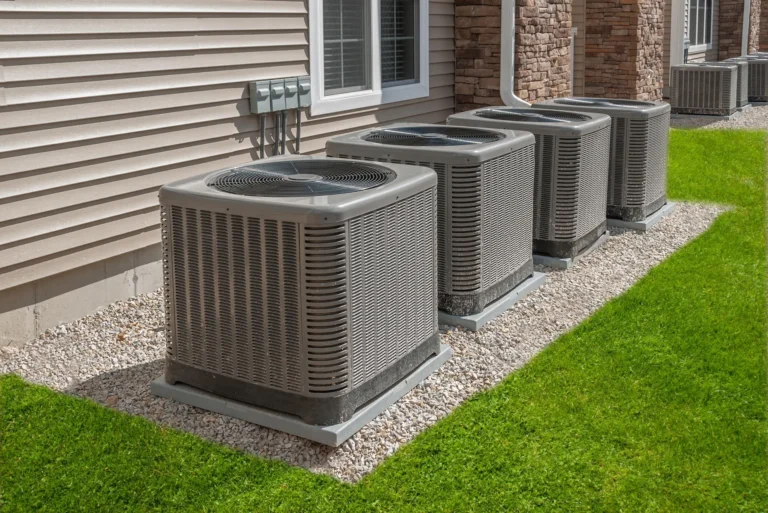 Four outdoor air conditioning units, expertly installed by Horizon Air, sit on a gravel bed beside a building with vinyl siding and brick accents, surrounded by lush green grass.