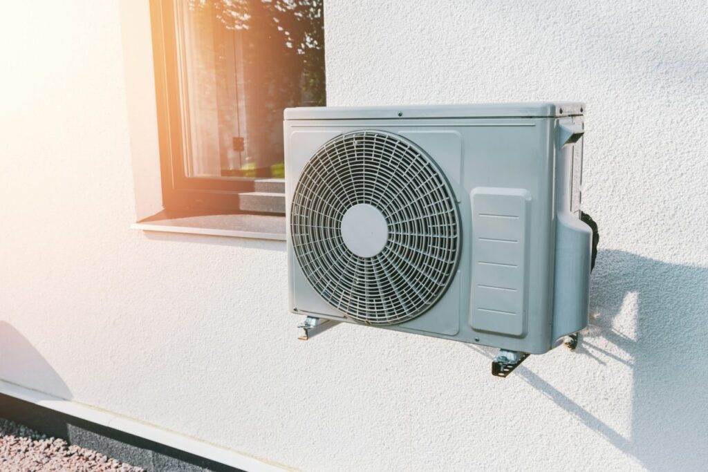 An outdoor air conditioning unit, installed during a professional AC Installation, is mounted on the exterior wall of a building next to a window on a sunny day.