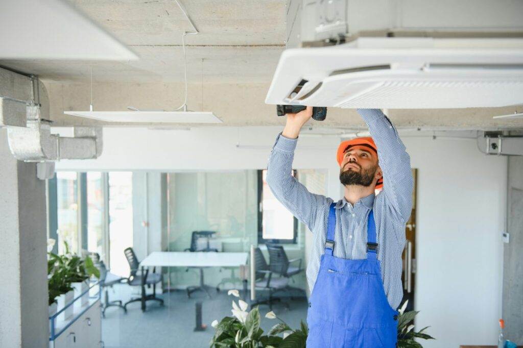 A worker in blue overalls and an orange helmet uses a power drill to service a ceiling-mounted air conditioning unit in an office, demonstrating the expertise of Horizon Air’s AC repair services.