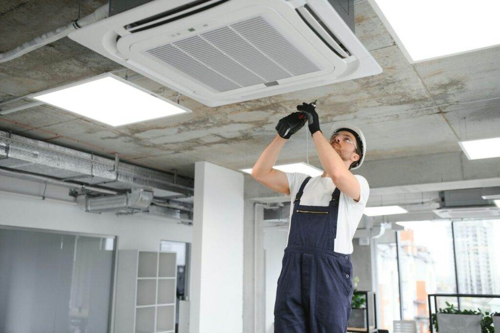 A worker in overalls and a hard hat is performing AC maintenance on a ceiling-mounted air conditioning unit in a modern office space.