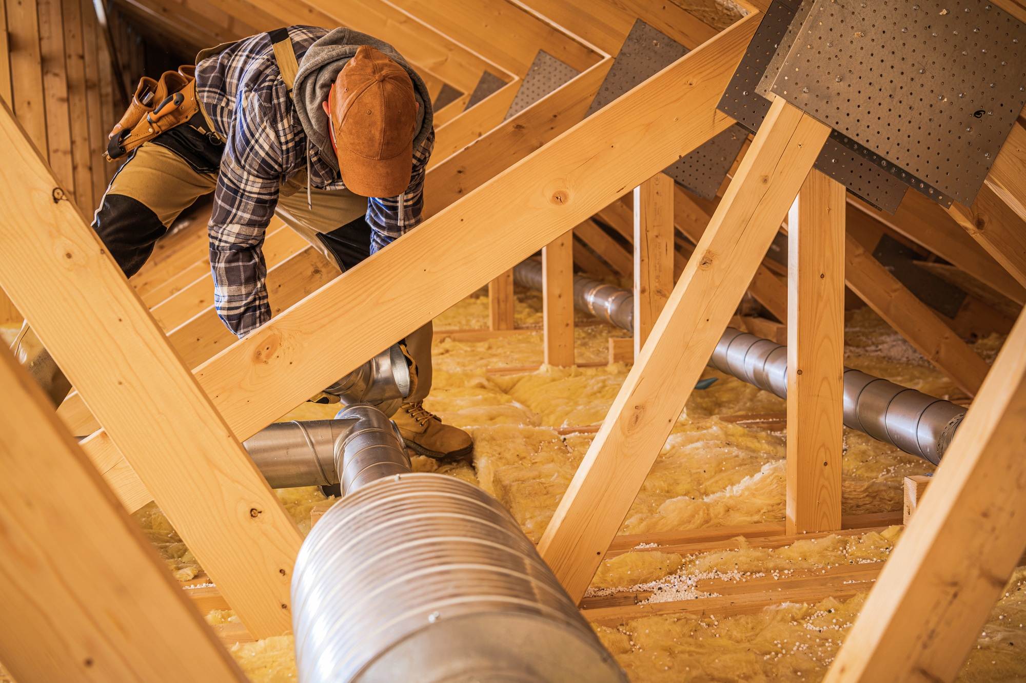 A worker in a flannel shirt and cap installs or inspects metal ductwork in a wooden attic with exposed insulation and beams, ensuring proper air conditioning and AC maintenance.