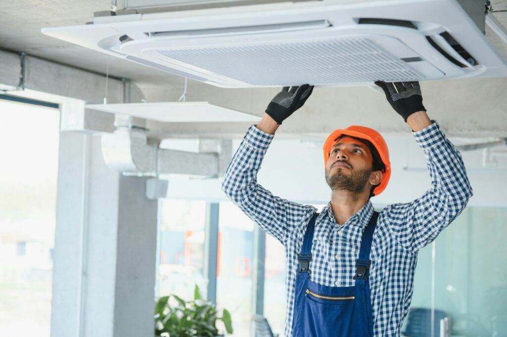 A worker in a hard hat and gloves inspects or repairs a ceiling-mounted air conditioning unit in a modern indoor setting, demonstrating Horizon Air’s commitment to expert AC installation and maintenance.
