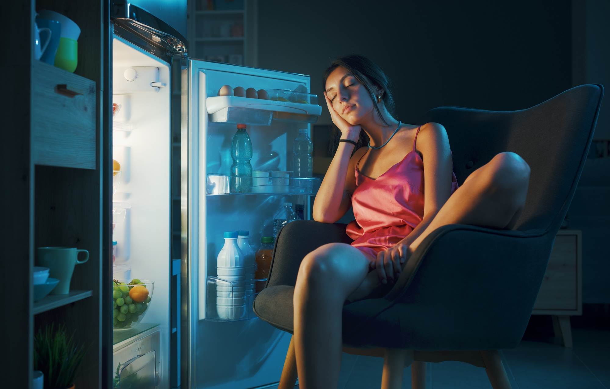 A woman in a pink satin dress sleeps in a chair next to an open refrigerator at night, illuminated by the fridge light—clearly needing some AC Repair or Maintenance for a better night's rest.