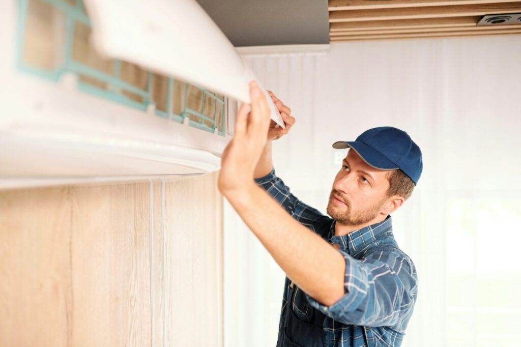 A man in a blue cap and plaid shirt inspects or performs AC Maintenance on an air conditioning unit mounted on a wall.